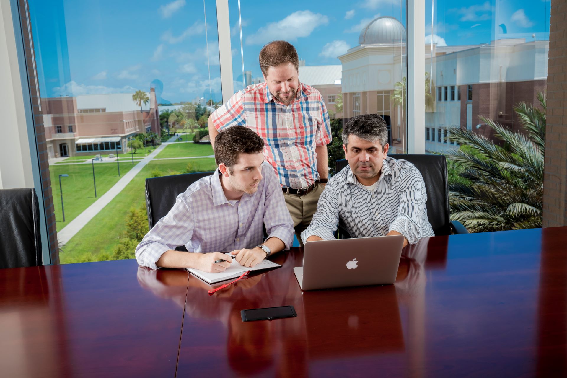 Three professionals in a modern office with large windows overlooking a campus, discussing work over a laptop at a glossy conference table